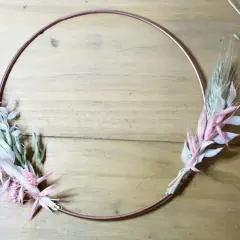 Étape d'un diy couronne fleurs séchées : un cercle de cuivre orné de bouquets de graminées et de fleurs séchées roses et blanches sur fond de bois.