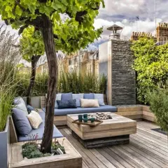 Aménagement terrasse devant maison : salon extérieur cosy avec banquette bleue, table basse en bois, arbres et végétation luxuriante.