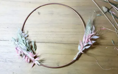 Étape d'un diy couronne fleurs séchées : un cercle de cuivre orné de bouquets de graminées et de fleurs séchées roses et blanches sur fond de bois.