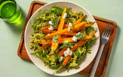 Pâtes au pesto avec des carottes rôties et des éclats de fromage de chèvre, garnies de fanes de carottes fraîches.