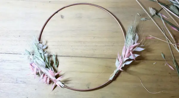 Étape d'un diy couronne fleurs séchées : un cercle de cuivre orné de bouquets de graminées et de fleurs séchées roses et blanches sur fond de bois.