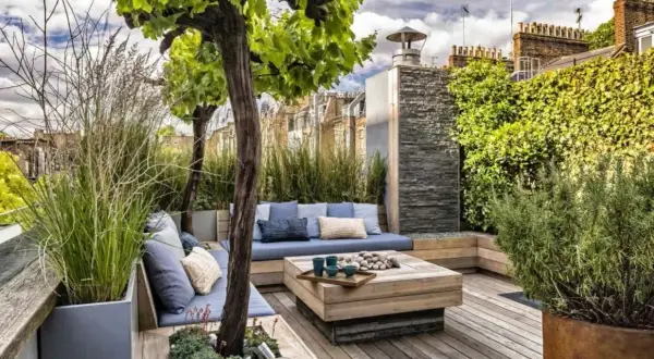 Aménagement terrasse devant maison : salon extérieur cosy avec banquette bleue, table basse en bois, arbres et végétation luxuriante.