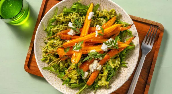 Pâtes au pesto avec des carottes rôties et des éclats de fromage de chèvre, garnies de fanes de carottes fraîches.