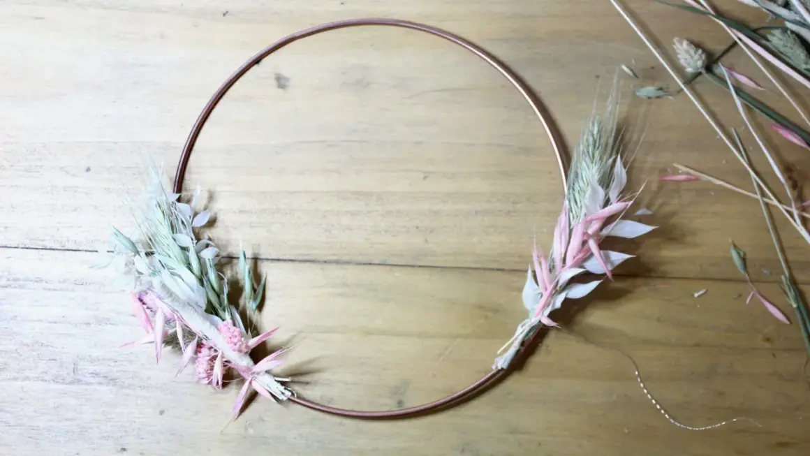 Étape d'un diy couronne fleurs séchées : un cercle de cuivre orné de bouquets de graminées et de fleurs séchées roses et blanches sur fond de bois.