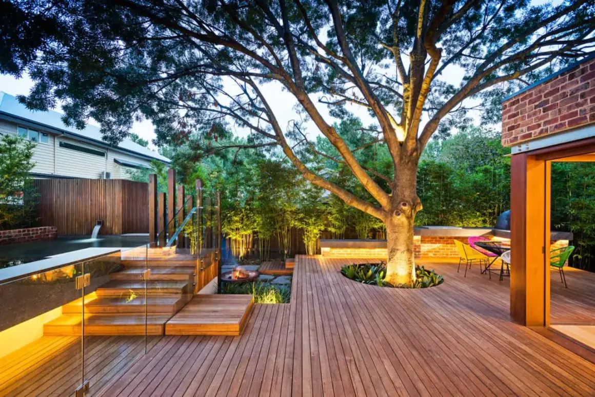 Un grand arbre est mis en valeur par un éclairage subtil, avec une **bordure autour de l'arbre** intégrée dans une terrasse en bois moderne.