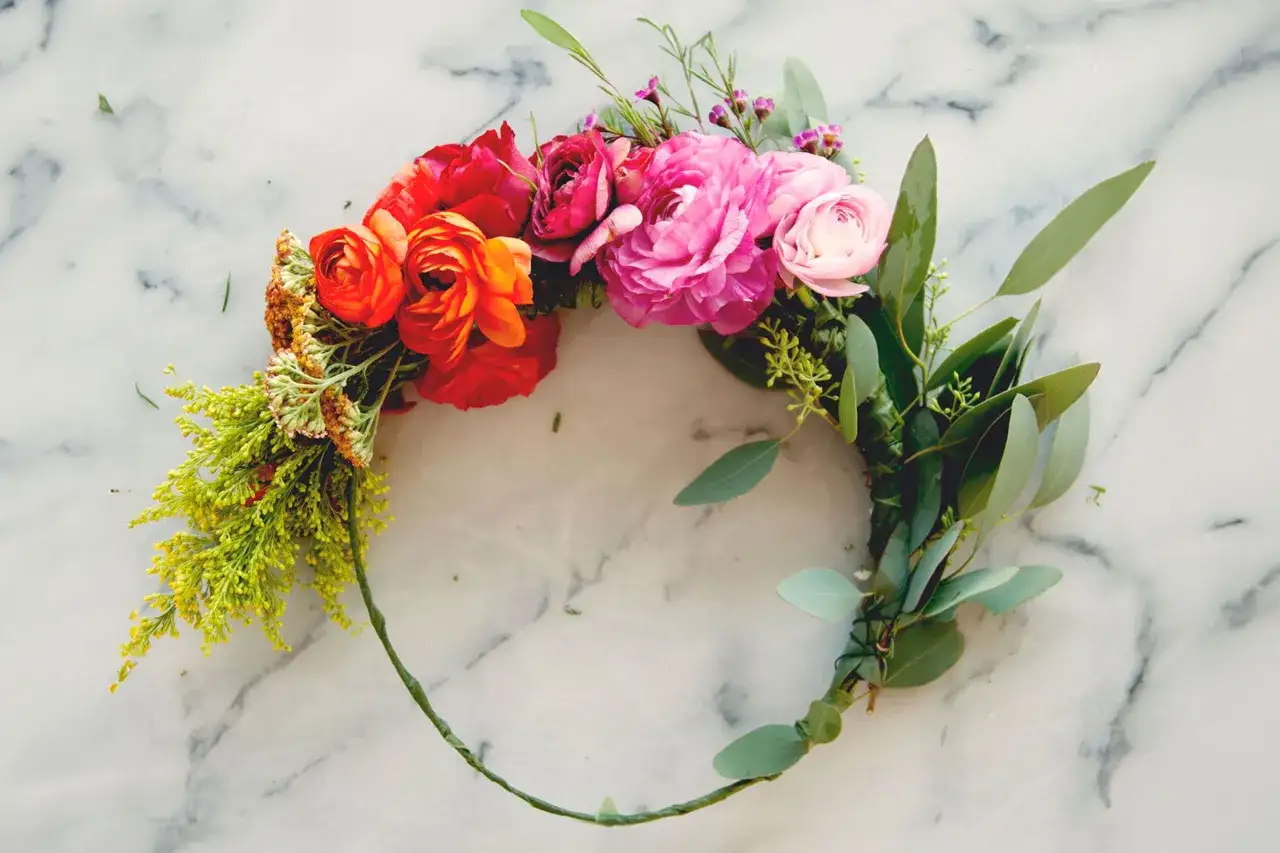 Une magnifique diy couronne de fleurs, ornée de roses rouges, roses et orange, de feuillage d'eucalyptus et de petites fleurs jaunes, sur fond marbre.